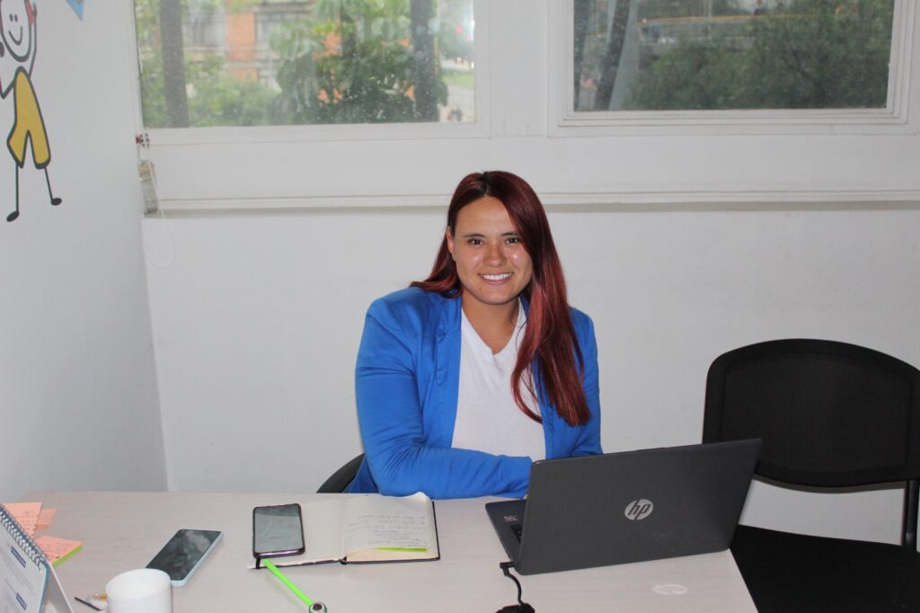 Mujer sentada en escritorio con blusa blanca, saco azul y cabello rojo sonriendo